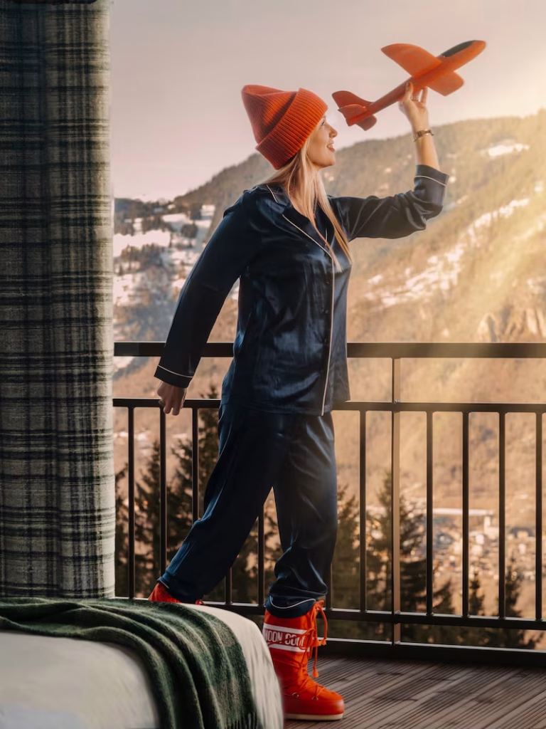 Femme en pyjama de soie bleu marine et bonnet rouge faisant voler un avion en jouet, balcon avec vue montagne.
