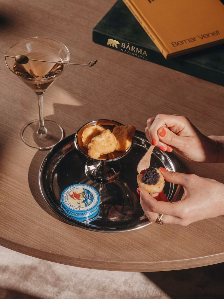 Gros plan sur un cocktail, des blinis et une boite de caviar, mains preparant une bouchee, livre Bârma en arriere-plan.