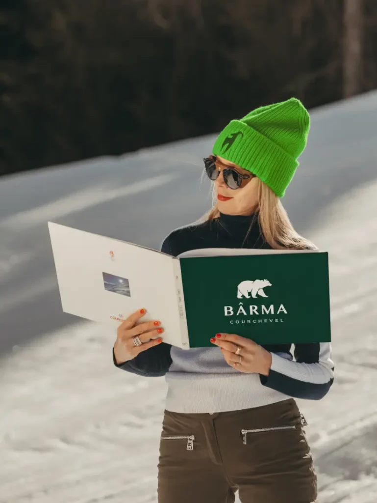Femme en bonnet vert lisant le livret de bienvenue Bârma Courchevel, piste enneigee en arriere-plan.