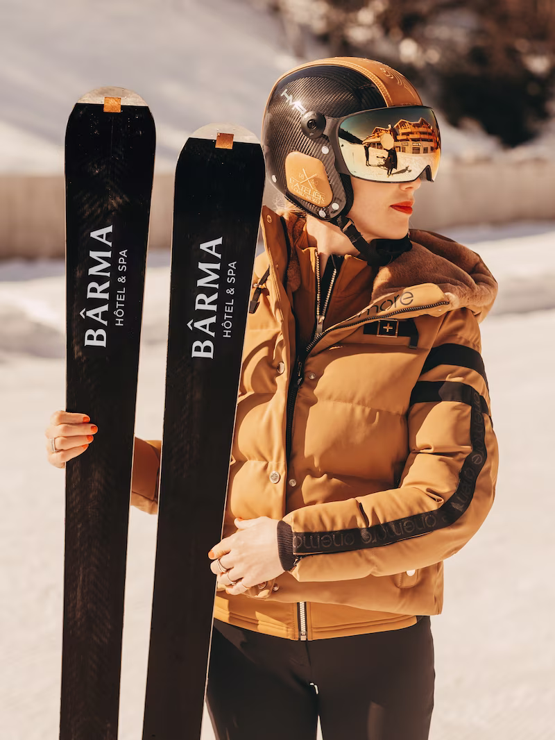 Femme casquee et lunettes de ski tenant deux paires de skis floques 'Bârma Hôtel & Spa', ambiance neige.