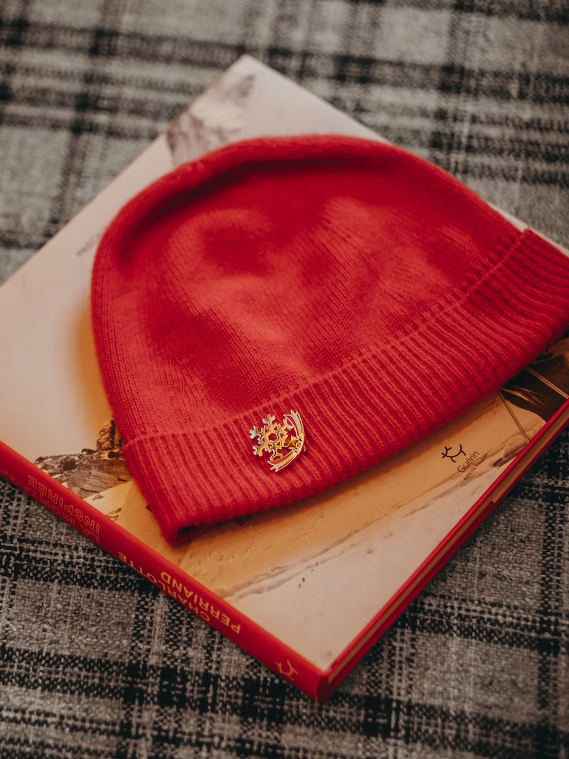 Bonnet rouge orne d'un pin's flocon-ski dore, pose sur un livre relie rouge, plaid a carreaux.