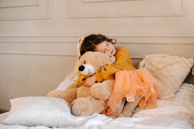 Petite fille en robe tutu orange serrant un grand ours en peluche dans ses bras sur un lit.