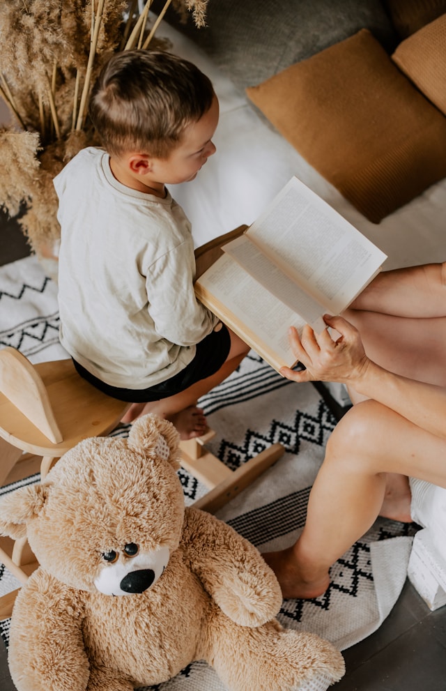Petit garcon assis sur un tabouret lisant un livre avec un adulte, ours en peluche au premier plan.