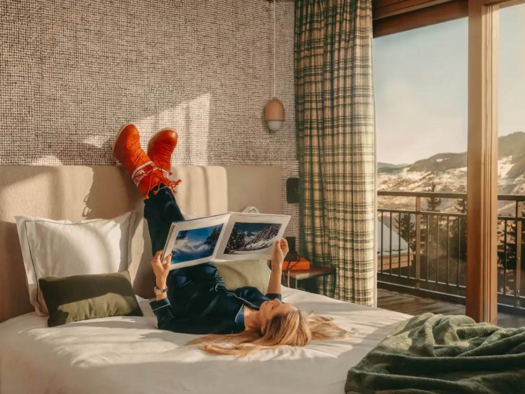 Femme allongee sur un lit, bottines orange sur la tete de lit, livre de photos en main, vue balcon sur le village enneige.
