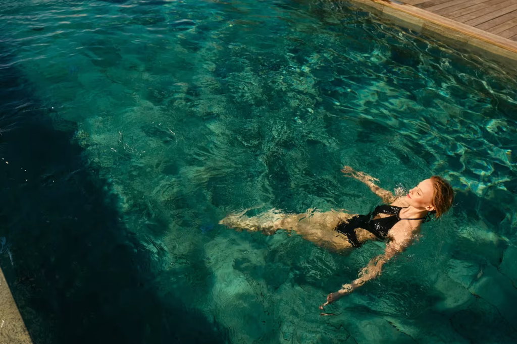 Femme flottant paisiblement dans une piscine aux eaux turquoise, maillot noir, lumiere du jour.