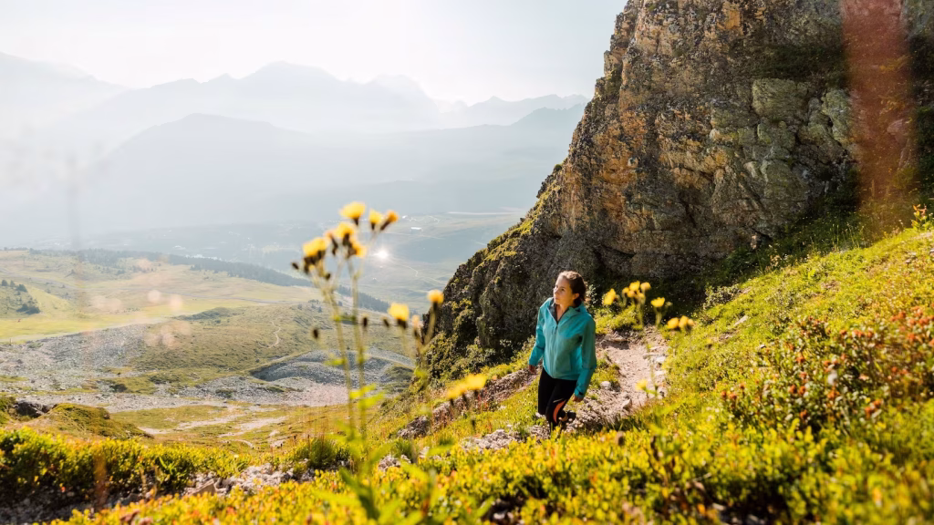 Femme en veste turquoise marchant sur un sentier rocheux, fleurs sauvages en premier plan.